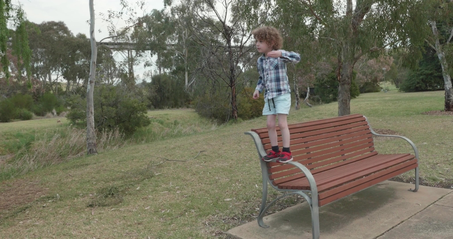 little boy jumps from the armrest of a park bench to the ground and then falls back on to his bottom 