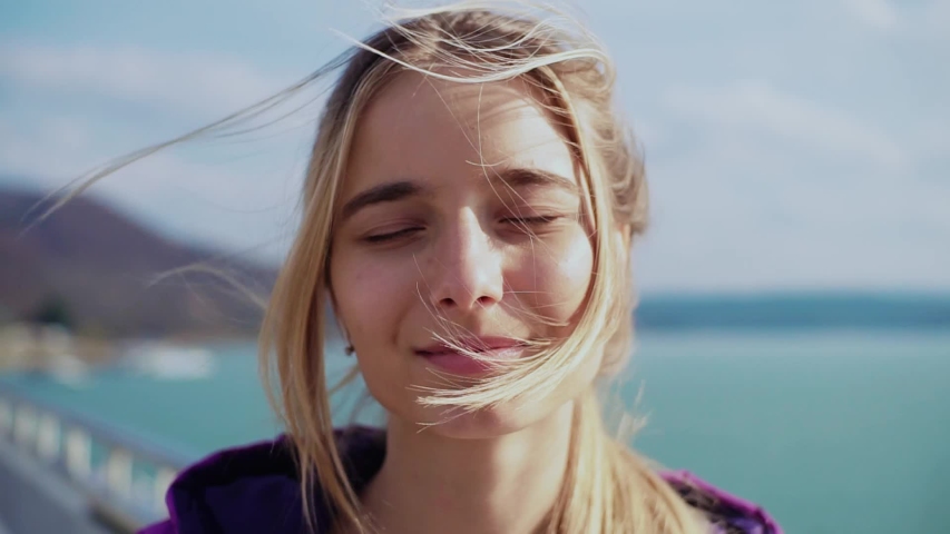 Happy face beautiful cute young woman look at camera wind shakes the hair on sunny urban sidewalk enjoying lifestyle trip natural outdoors pretty portrait close up slow motion