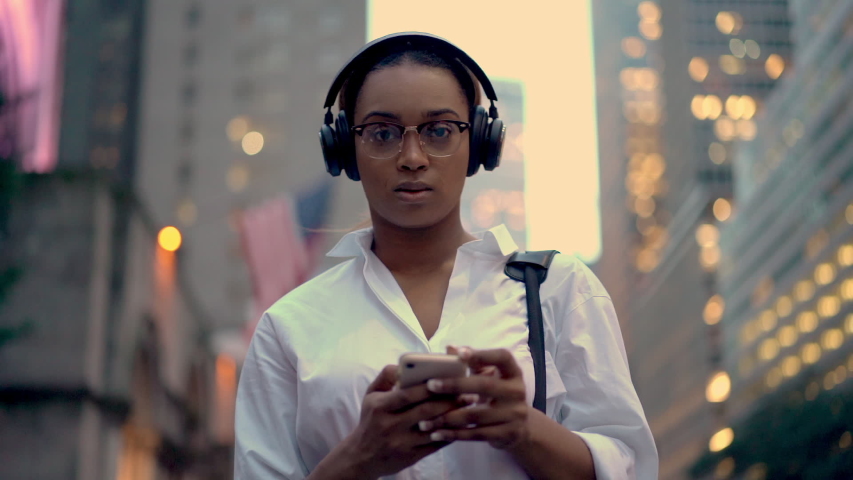 Slow motion of business consultant in stylish spectacles standing at urban street and using banking application on smartphone gadegt for making online money transfer, headphones for noice cancellation