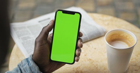 Lviv, Ukraine - August 09 , 2019: Hands of Afro American Man Sitting at Table with News Paper and Coffee Cup on it, Holding Smartphone with Green Screen and Drinking Coffee. Backside View. Close Up