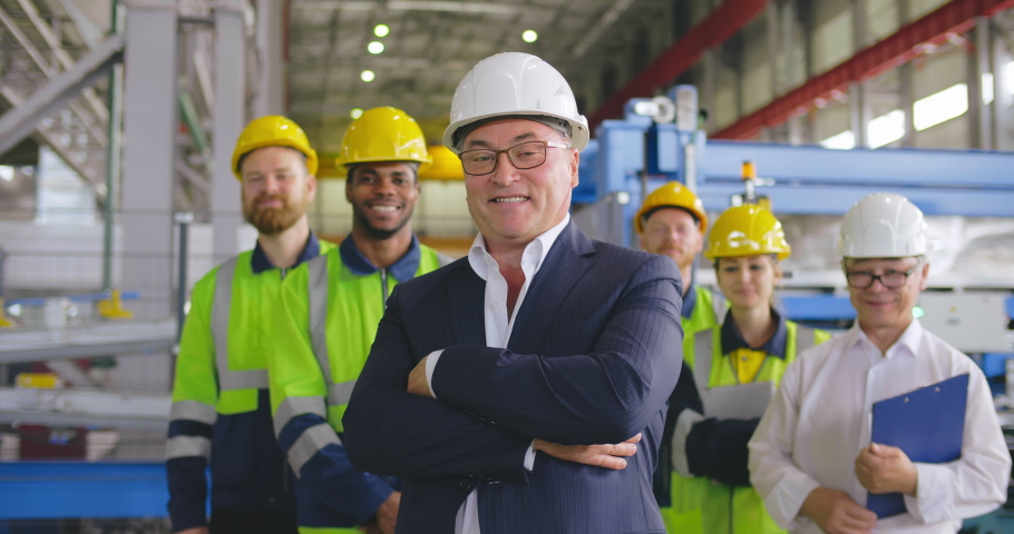 Caucasian businessman smile face portrait. Positive smiling business person in formal wear, helmet looking to camera crosses arms over chest. On background staff team employees at heavy industry plant Royalty-Free Stock Footage #1042494667