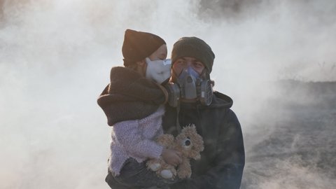 A survivor father carring her daughter in gas mask going through clouds of toxic smoke in empty dead landscape.