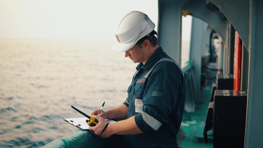 Marine chief officer or chief mate on deck of ship or vessel. He fills up ahts vessel checklist. Ship routine paperwork. He holds VHF walkie-talkie radio in hands. Royalty-Free Stock Footage #1042502818