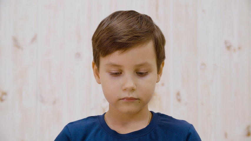 Portrait of adorable pensive boy looking down and winking. Close-up view of cute serious preteen boy standing and looking down indoors