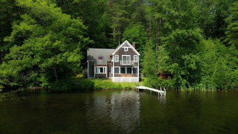 Secluded cottage in the woods on a pond with a dock and tall trees surrounding the peaceful scene in the forest. Drone flies closer.