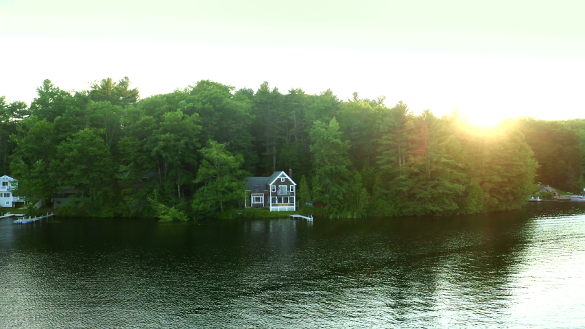 DRONE - Vacation house cabin on a lake in New England with a pond and blue water dock. Camera flies towards the scenic house on the water with sunset filtering through the trees.