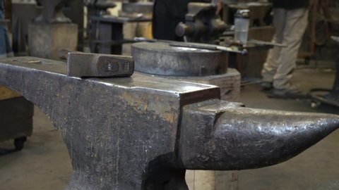 We can see a hammer on an anvil in a blacksmith factory. The blacksmiths are moving a lot of behind the anvil.