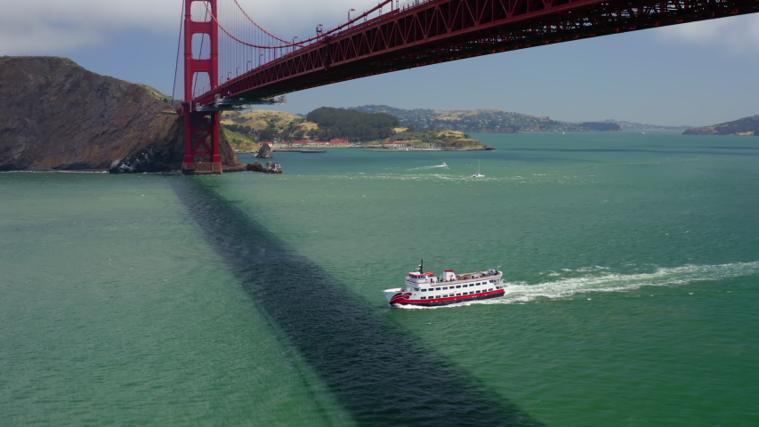 Aerial 4K footage of red and white passenger ship passing under the beautiful and well-known Golden Gate Bridge. The panorama of the coast is at the background. Bridge shadow is seen on the water.