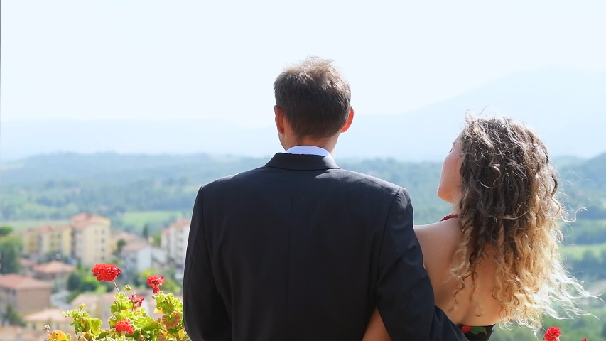 Outdoors balcony terrace garden in Italy with aerial high angle view of rolling hills in Chiusi, Tuscany young elegant woman and man couple with hair flying in wind, kissing each other