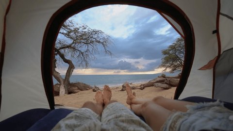 Young couple lying in a camp tent closed up point of view on male and female legs with sea view on stunning beach at sunset 