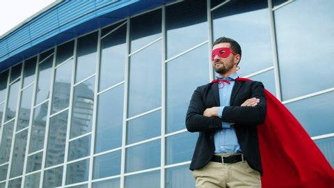 Portrait of handsome young businessman in suit wearing superman costume outdoors standing alone near office building, red cape is wavering in the wind.