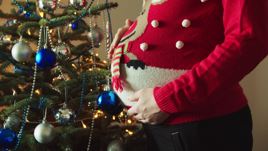 Young 9 months pregnant mother cuddling her tummy in the Christmas sweater in front of the decorated Christmas three
