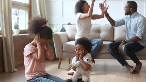 Afro frightened little kids sit on floor covered ears not to hear parents quarrel, couple sorts out relations on sofa in presence of children, negative situation affects childs mental health concept