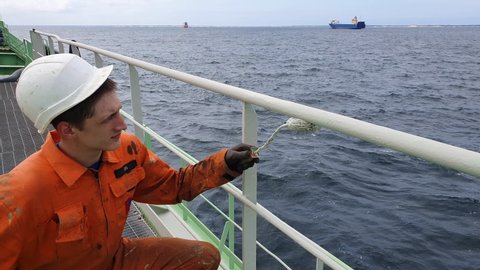 ship worker in helmet sits on haunches and paints railings in white against calm dark blue sea with tanker on raid closeup