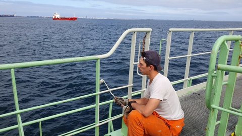 sailor in rubber gloves sits on haunches and paints railings white color against calm dark blue sea with red tanker