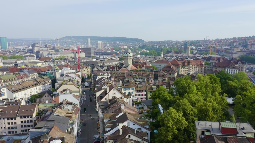 Zurich, Switzerland. An aerial view sweeps across the historic old town, capturing the distinctive 51-meter octagonal concrete tower of the Urania Sternwarte Observatory rising above dense clusters of traditional European rooftops with warm brown tiles. The camera glides over Lindenhof hill, a heritage-listed park occupying a moraine hill that has served as a strategic settlement site since Roman times, its lush green canopy contrasting with the stone and plaster architecture below. The Limmat River valley stretches toward the horizon, framed by rolling hills under a bright summer sky. The scene blends centuries of urban development with vibrant natural greenery, presenting a sweeping panorama of one of Europe