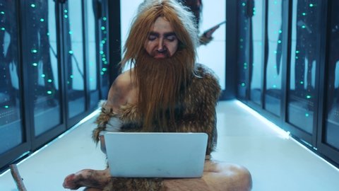Gloomy prehistoric human in animal fur discovering technology in database server. Ancient IT engineer typing on laptop winning celebrating business success working at data center.