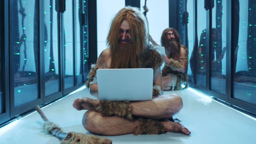 Euphoric caveman celebrating business success using a laptop clenching fists rejoicing while sitting on floor in server cabinet. Prehistoric happy IT specialists working at data center. Royalty-Free Stock Footage #1042548628