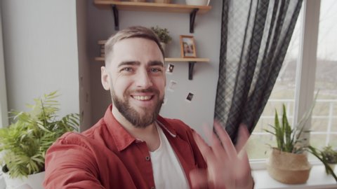 Chest-up handheld shot of happy young Caucasian man with invisible smartphone in outstretched hand standing by window at home and having lively conversation with someone on video call