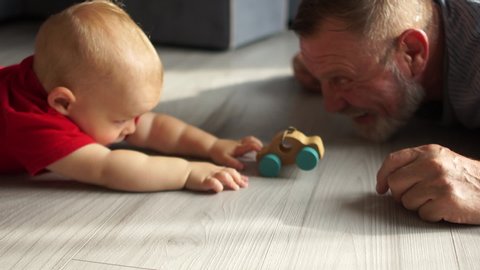 Grandfather playing with little grandson with a toy wooden car, caring granddad and cute preschool grandchild lying on warm floor together, having fun at home, underfloor heating concept close up