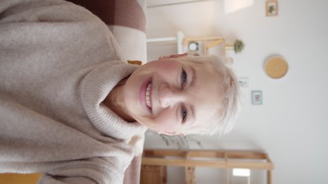 Vertical handheld waist-up shot of senior Caucasian woman with short grey hair sitting on couch at home, holding invisible smartphone in outstretched hand and chatting with someone on video call