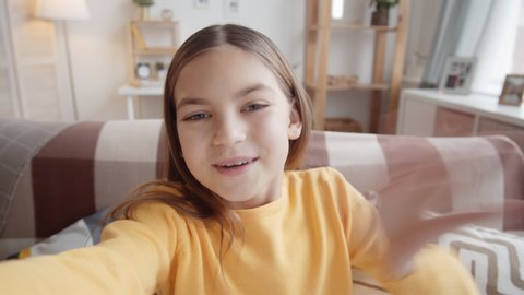 Chest-up handheld shot of happy 11-year-old Caucasian girl sitting on couch at home, holding invisible smartphone in outstretched hand, looking at camera and chatting cheerfully on video call