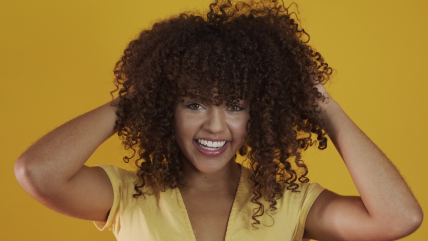 Happy laughing American African woman with her curly hair on yellow background. Laughing curly woman in sweater touching her hair and looking at the camera. Royalty-Free Stock Footage #1042561081