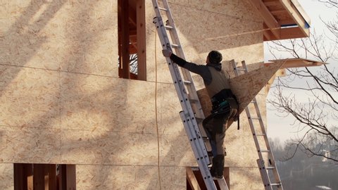 Professional builder is climbing high steel ladder with triangle plywood. Frame residential house under construction
