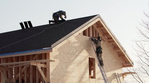 Residential house under construction. One worker is covering the wooden frame with plywood using hydraulic hammer, another one is making ridge on the roof in safety harness