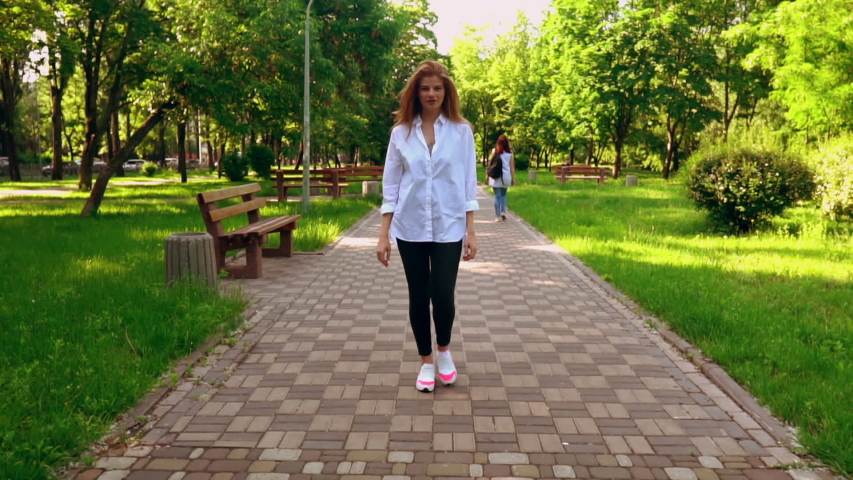 beautiful young woman walking to the camera cheerful smiling outdoors person with long red hair in white shirt walking in summer city green nature slow motion