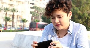 Portrait of a relaxed teenage boy sitting at the city park with smart phone. Kid watching to screen, reading, typing, playing games. Technology, internet communication and people concept