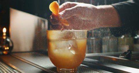 Close up of a professional bartender is squeezing an orange while preparing an alcoholic cocktail with ice cubes to customers at the bar or disco club.