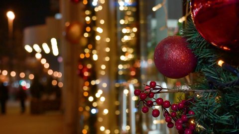 Winter evening Close up: store Doors are decorated with Christmas garland: 2 red balls and branch of berries Lights flash Blurred background: Caucasian woman in black down jacket walks out door