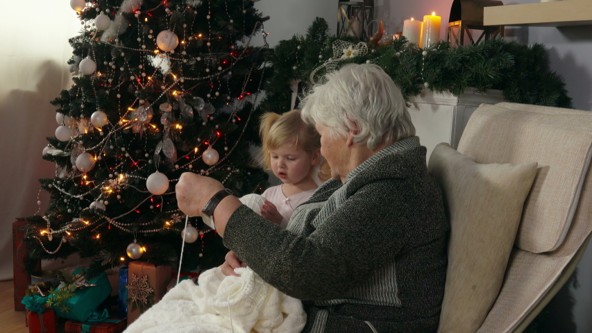 Elderly woman knits white blanket sitting in a chair with a little cute girl in pink dress next to the Christmas tree. Grandmother shows to her granddaughter how to knit