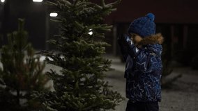 beautiful slow motion video - little boy fun shakes off snow from a snowy Christmas tree in the park on a winter evening