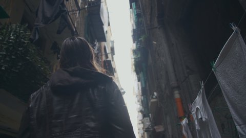 Brown haired girl walks between tall decrepit buildings in an unlit alleyway in Europe. Young woman in her thirties walking alone in an old neighborhood alleyway. Slow-motion, back view, side view.