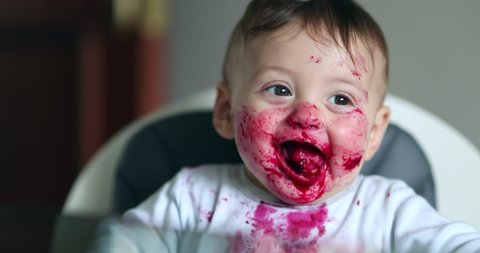 
Adorable baby covered with red sauce. Feeding messy infant on high-chair