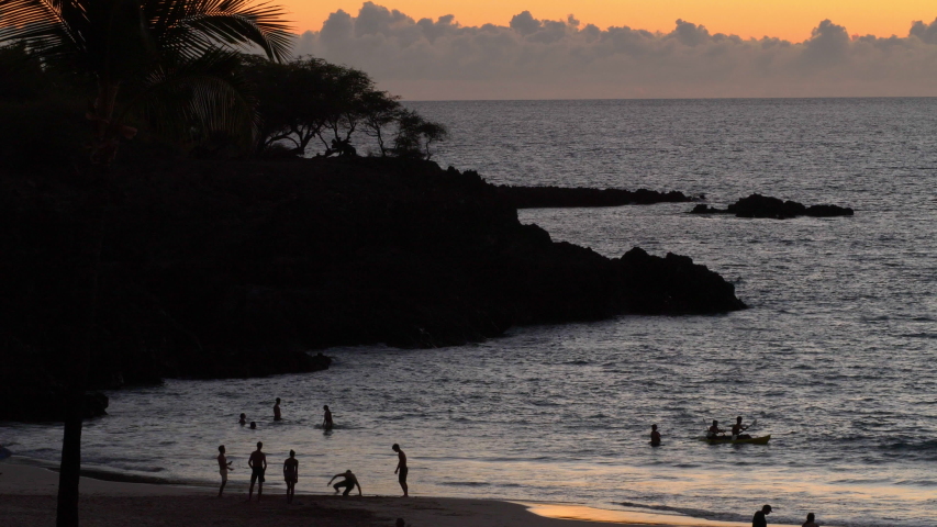 A Hawaii beach at sunset.  Distant silhouettes of people play on the beach and paddle in a boat