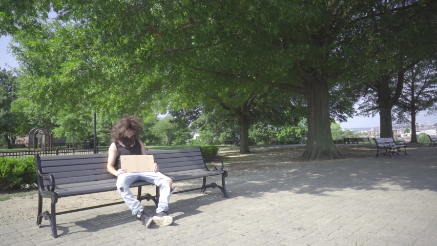 Bearded homeless man with sign sleeps on bench in park, summer day