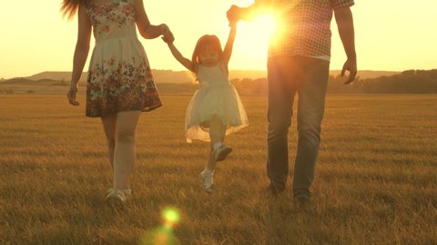 little daughter jumping holding hands of dad and mom in park on background of sun. Family concept. child plays with dad and mom on field in sunset light. Walking with small kid in nature.