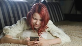 teenager in sweater lies at home on sofa on pillow with smartphone. girl with the phone reads the message and smiles cheerfully. Chat with friends and family. View photos and videos in modern gadgets.