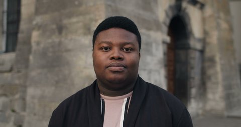 Close up of happy fat man looking to camera. Young african guy standing outdoors and smiling. Portraits. Daylight. Concept of people and emotions. Old buildings background