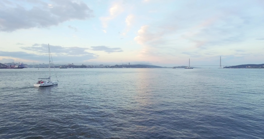 Aerial view of bautiful white sailing yacht with high mast in the Eastern Bosphorus strait at summer sunset. Vladivostok cityscape and Russian bridge are on the background