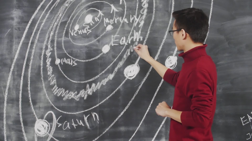 Lockdown of young Asian man standing at blackboard and drawing universe map on it using piece of chalk