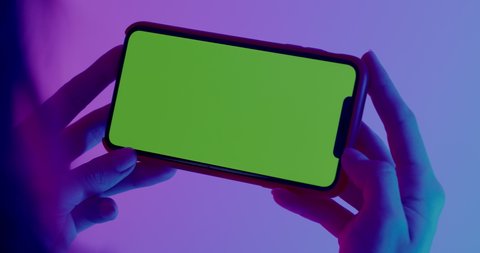 MINSK/BELARUS - CIRCA 2019: GREEN SCREEN CU Caucasian female using Apple iPhone XS Max, horizontal orientation, colorful neon background. 4K UHD RAW Graded footage