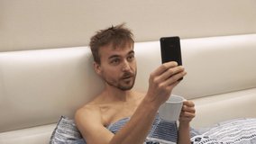 Young male blogger uses smartphone lying in bed with cup of coffee in his hand. Millennial man records post for his followers early in the morning. Bearded guy video chatting in bedroom.