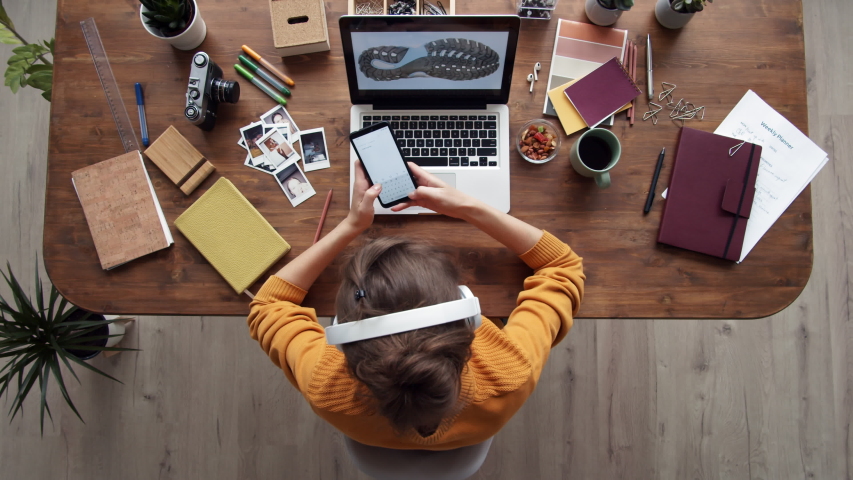 Time lapse of young woman sitting at laptop, putting earphones on head while looking through photos of footwear on PC, using phone, writing in notebook and drinking tea