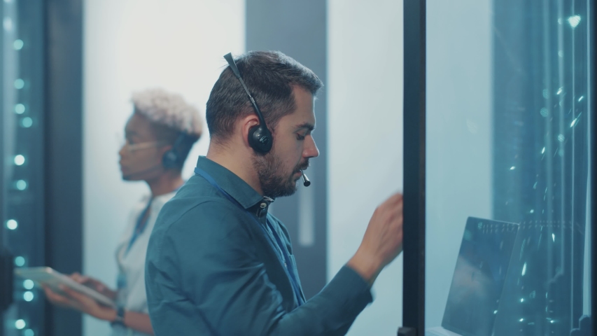 IT support engineer hosting on laptop computer at server racks, talking with head office on headphones, coworking in team at database cyber security.