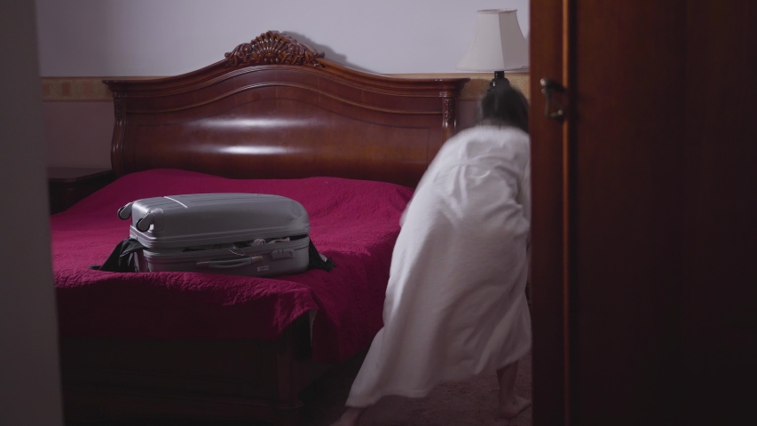 Portrait of young Caucasian woman closing travel bag with clothes and running out the room.  Lady in white bathrobe gathering stuff before a trip.