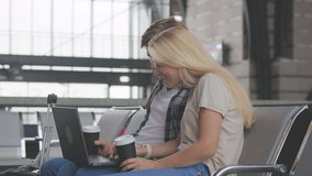 Cheerful woman holding paper cup near boyfriend with laptop while sitting in airport waiting hall. Sweet couple booking train tickets for their travel. watching fun videos by internet online